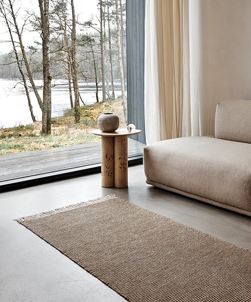 A serene living room interior featuring the Pappelina Lina linen rug, placed on a polished concrete floor. The room includes a beige sofa, a wooden side table with a ceramic vase, and large windows offering a view of trees and a lake.